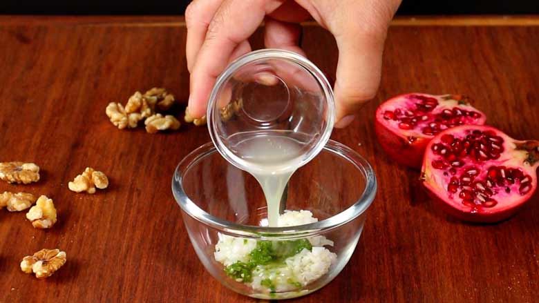 A hand is seen pouring lime juice from a small glass bowl into another glass bowl with onion and green chillies to make a salad dressing. California walnuts kernels and an open pomegranate lie on the wooden counter in the background - Konkani Cucumber and Pomegranate Salad Recipe
